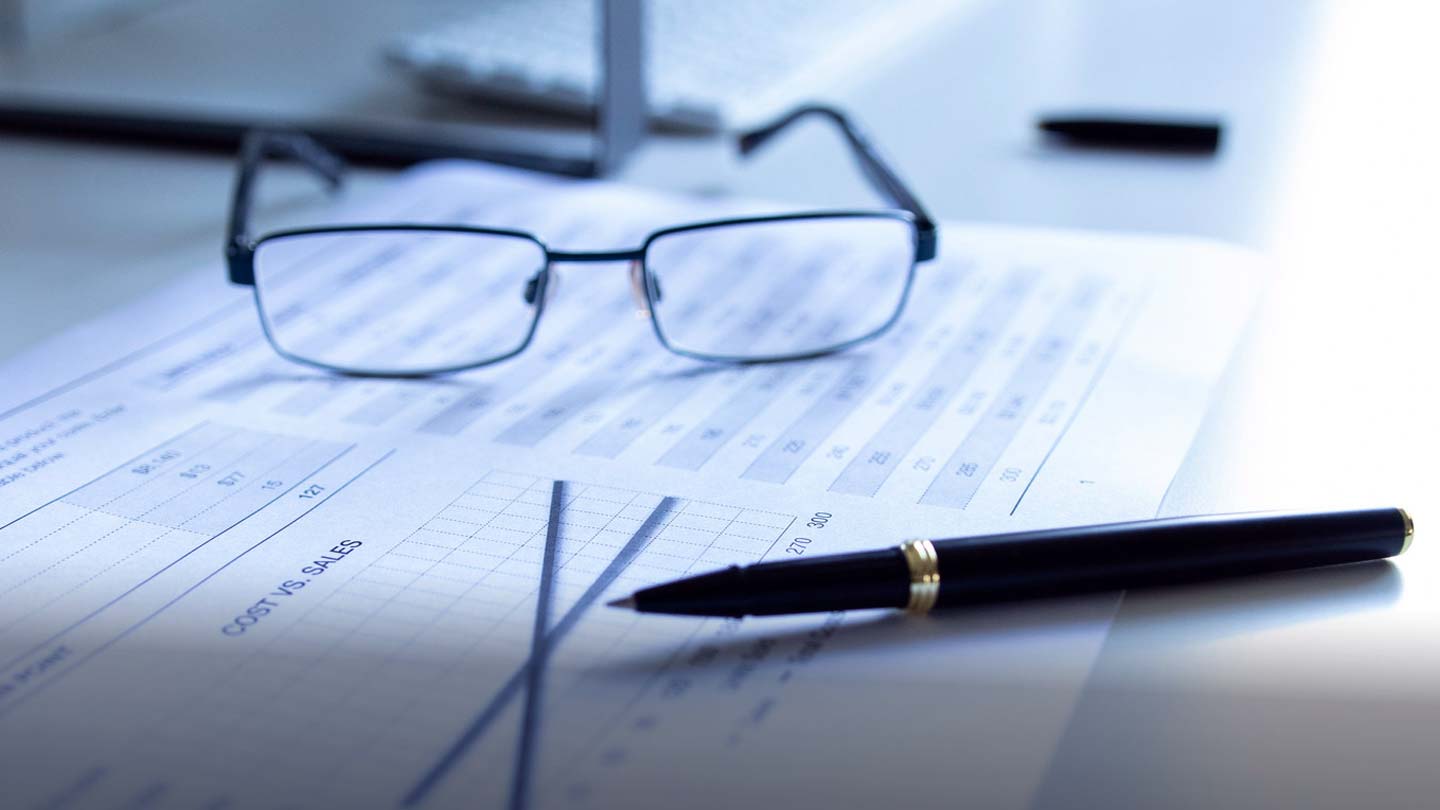 A close-up of glasses and a pen on a sheet of paper with printed tables and graphs, indicating data analysis or financial review in an office or study environment.