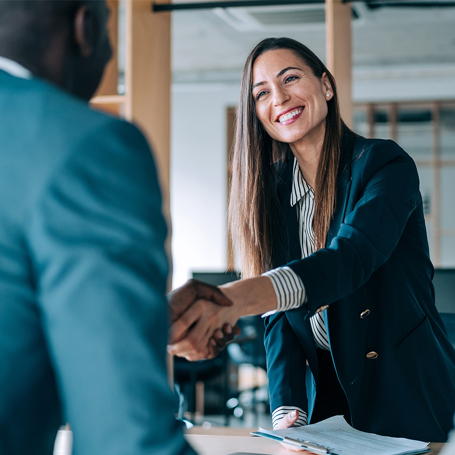 Two individuals shaking hands across a table in a professional setting, symbolizing agreement, introduction, or deal completion, with documents suggesting a business meeting.