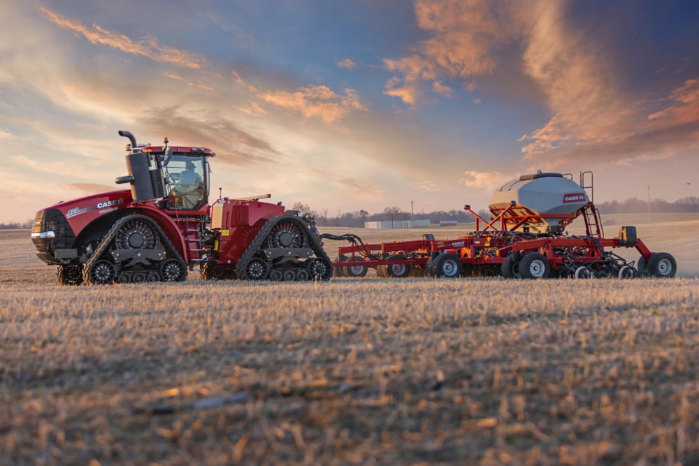 A large red Case IH tractor with tank treads pulls agricultural equipment across a field during sunrise or sunset. The warm hues of the sky blend into cooler tones, capturing the essence of a day’s work in farming.